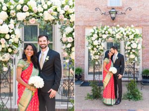 Man and woman posing for wedding photos. The bride wore a red traditional Indian sari and the man wore a black tuxedo. They posed under a large white and pink floral arch during their intimate summer backyard wedding.