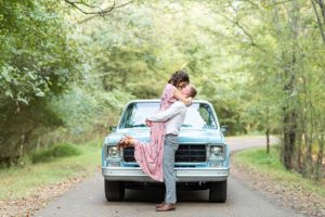 Man and woman kissing while the man lifts the woman off the ground in front of a vintage blue, Chevy truck