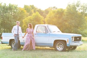 Photo of man and woman holding hands in a lush, green field in front of a vintage blue Chevy truck during anniversary session