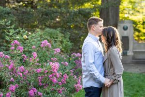 Couple posing near blooming flowers at Northwestern University engagement photo session. Man and woman are holding hands while facing each other. The man is kissing the woman on the forehead.