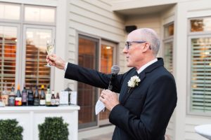 photo of an older man in a tuxedo giving a toast. he is holding a glass of champagne and a microphone.