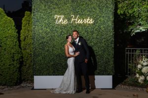 Bride and groom posing in front of a wall of greenery and a neon sign with their last name